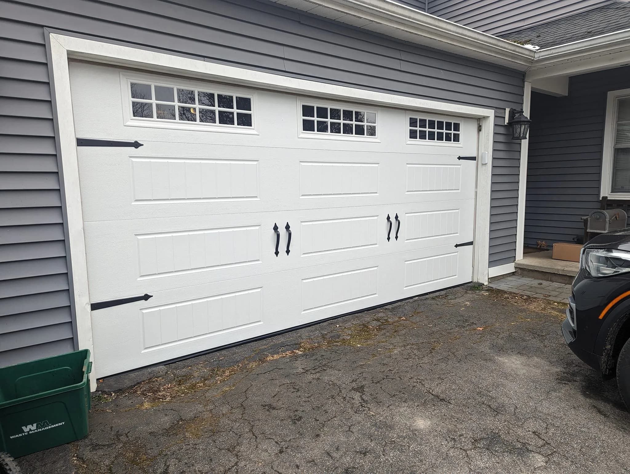 White carriage-style garage door with grid windows and iron hardware on a gray-sided home