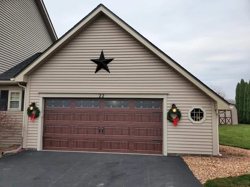 Walnut woodgrain carriage-style garage door with grid windows and holiday wreaths on a tan-sided home