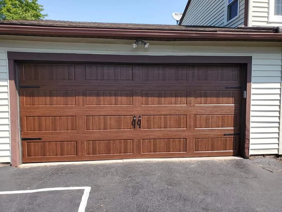 Walnut woodgrain carriage-style garage door with iron hardware on a home with blue-green siding