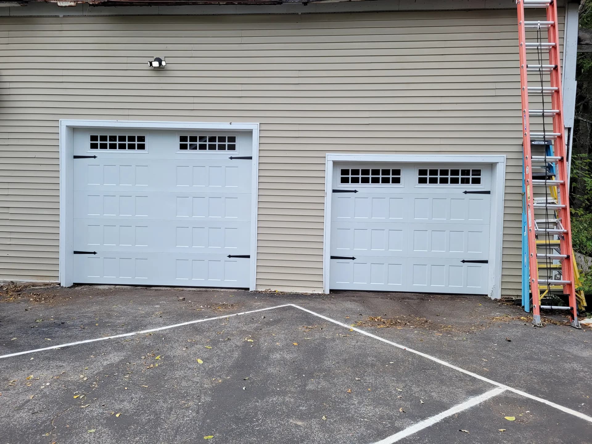 Two white carriage-style garage doors with grid windows and iron hardware on a tan-sided home