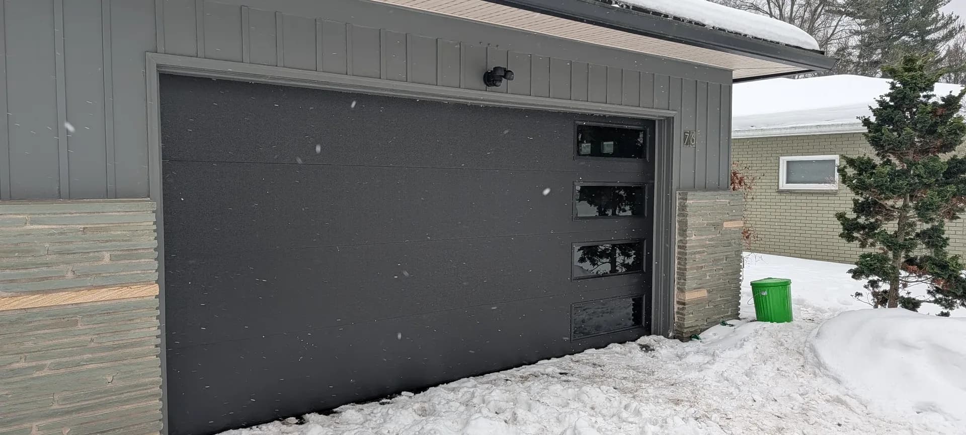 Charcoal raised-panel garage door with vertical windows on a ranch home with green siding in winter